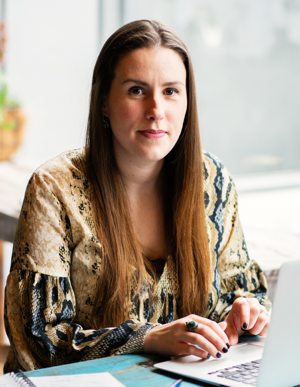 Professional with long brown hair wearing a patterned blouse sits at desk near window, reviewing documents in bright office setting