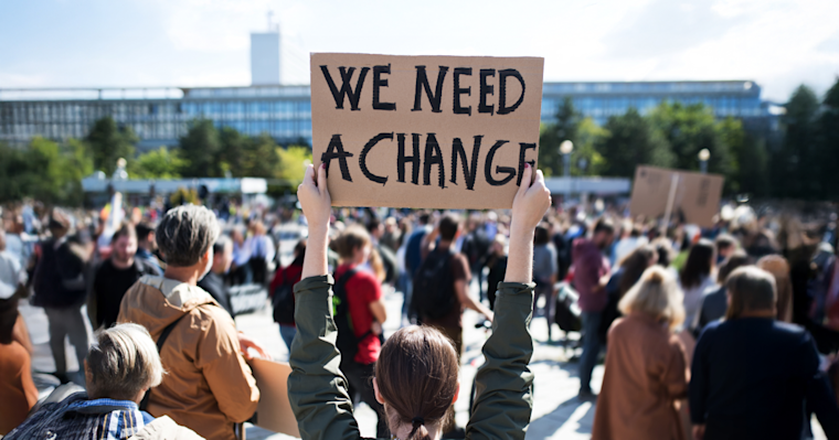 A person seen from behind holds a cardboard sign reading “We need a change” above their head at a large outdoor protest. A crowd of people gathers in a public plaza with buildings visible in the background. 