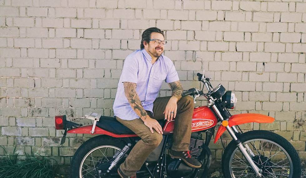 Person with tattoos wearing glasses and light blue shirt sits on vintage red Suzuki motorcycle against white brick wall