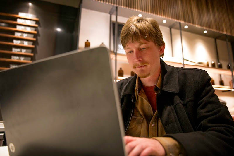 A person works on a laptop in a warmly lit modern space with wooden shelving and decorative bottles displayed in the background.