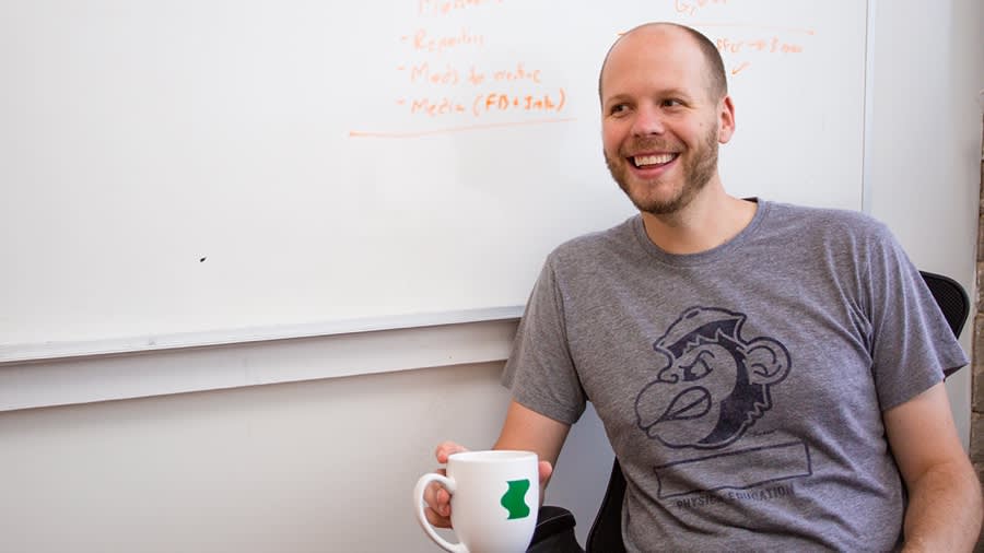 Smiling instructor in grey t-shirt with monkey graphic holds white mug near whiteboard with mathematical formulas written in pink