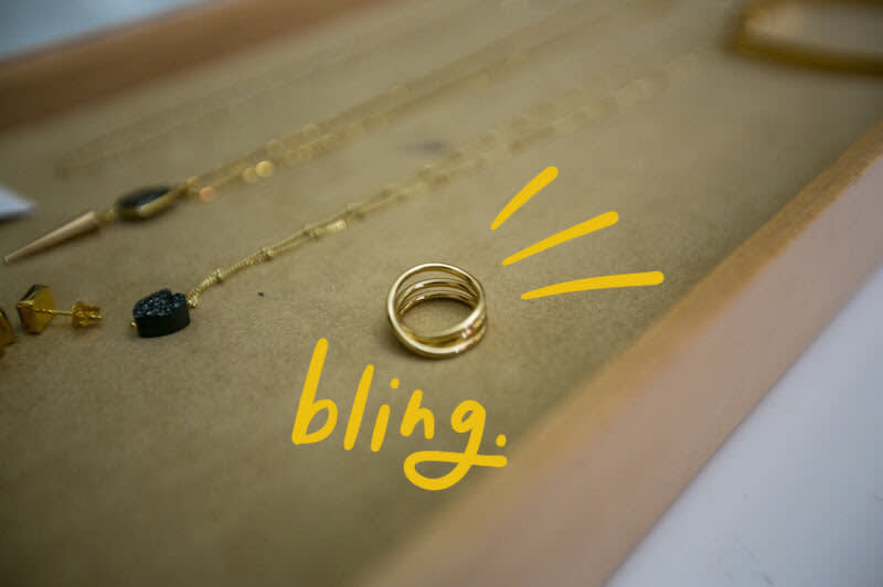 Gold ring on beige surface with hand-drawn yellow "bling" text and sparkle lines, jewelry pieces visible in background