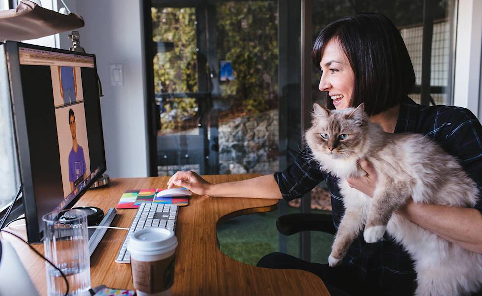 Woman in dark shirt holds fluffy Ragdoll cat while working at desk during video call, coffee cup nearby