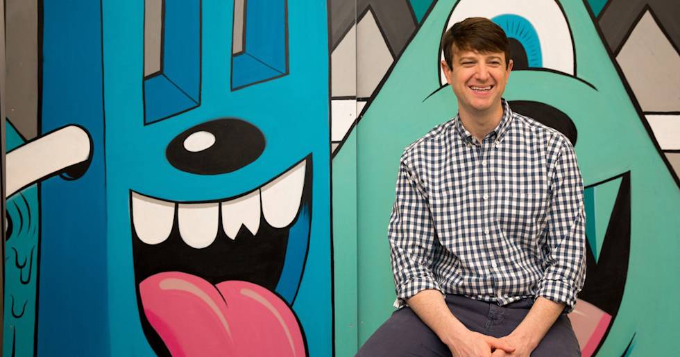 A person in a checkered shirt sits smiling against a vibrant mural wall featuring cartoon monster faces in blue and mint green colors.