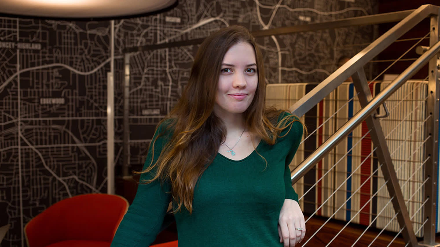 A person in an emerald green sweater stands by modern metal stairs, with a stylized map pattern visible on the dark wall behind them.