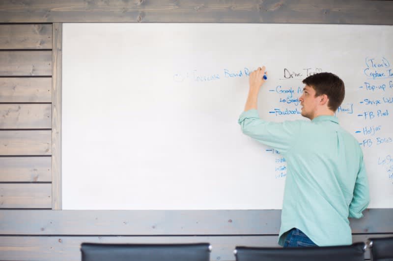 Person in mint green sweater writing on large white board mounted on wooden wall, viewed from behind