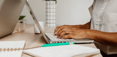 Person typing on laptop keyboard while working at white desk with natural lighting, cropped view showing hands