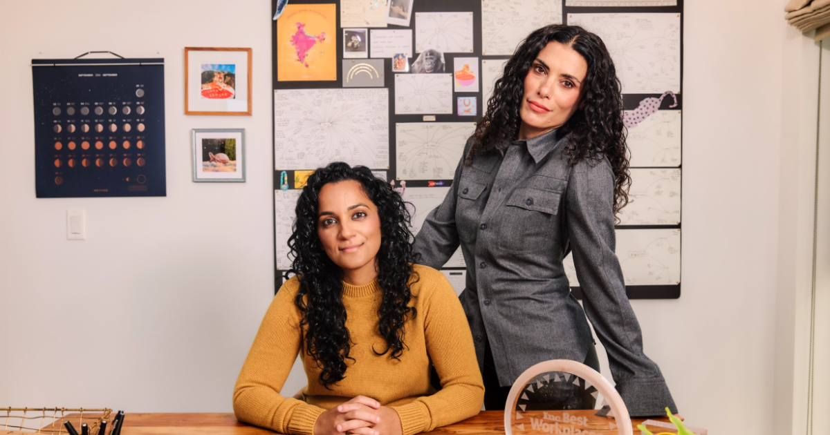 The CHANI co-founders pose in their office. Sonya Passi sits at the desk, and Chani Nicholas stands behind her with her hand on the back of the chair. In the center of the desk is a "2024 Inc. Best Workplaces" award.