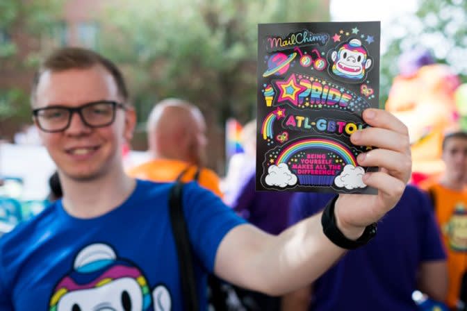 Person holding up colorful Pride-themed MailChimp sticker with rainbow designs and ATL-GBTQ text at outdoor event