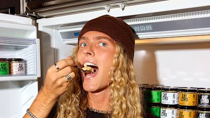 Person with long blonde curly hair and brown beanie eating from a spoon while standing in front of an open refrigerator stocked with Oat Lord products