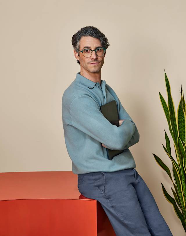 A man with glasses and a blue sweater sitting on a red table with his arms crossed, holding a laptop.