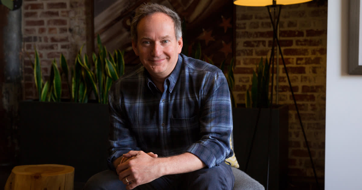 Professional in a blue plaid button-down shirt sits casually on a chair in a warm, brick-walled room with indoor plants