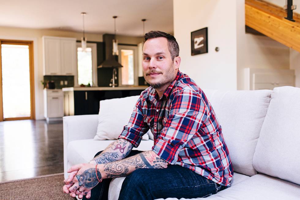 Person in plaid shirt with tattoos sitting casually on white couch in modern open-concept living space with wooden accents