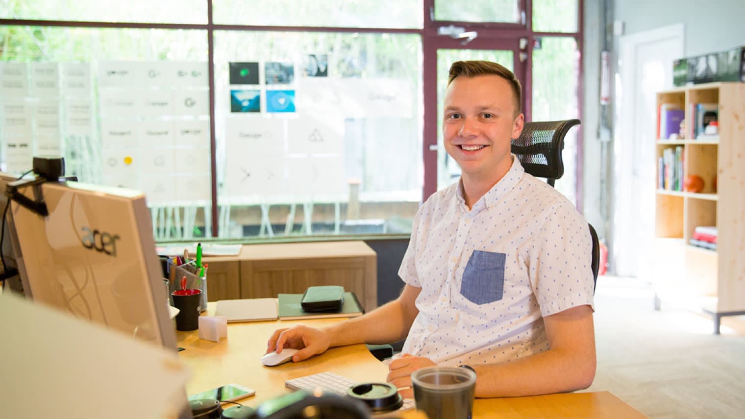 Professional working at modern office desk with Acer monitor, wearing white patterned shirt in bright workspace with whiteboard behind