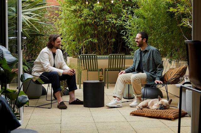 Two men having a cheerful conversation while seated seated on a patio decorated with plants and modern furniture