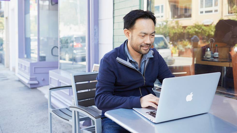 Man in navy jacket smiling while working on MacBook laptop at outdoor cafe table