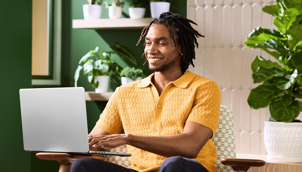 A man with locs smiles as he types on a silver laptop. He is wearing a textured yellow collared shirt and is positioned in a modern chair. Behind him, shelves and walls display various green house plants.