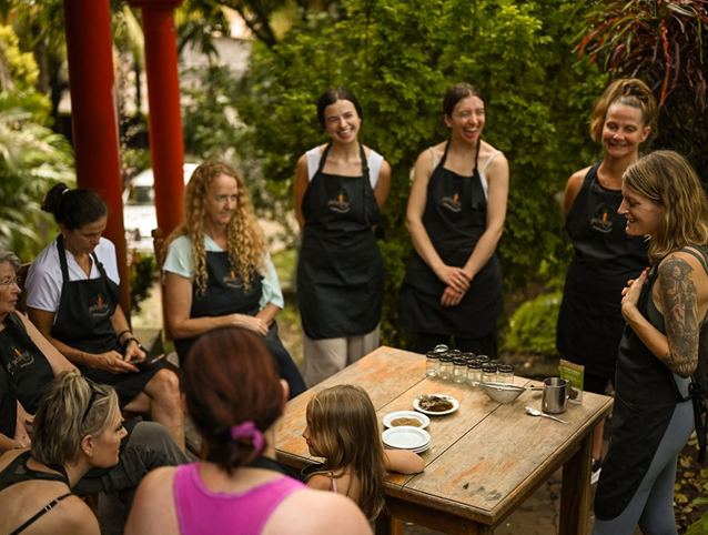 A group of people in black aprons gather around a wooden table during an outdoor cooking class or demonstration in a garden setting
