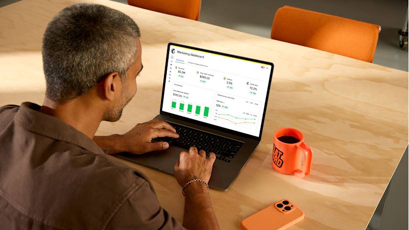 Professional reviewing marketing dashboard on laptop at wooden desk with orange coffee mug and phone nearby