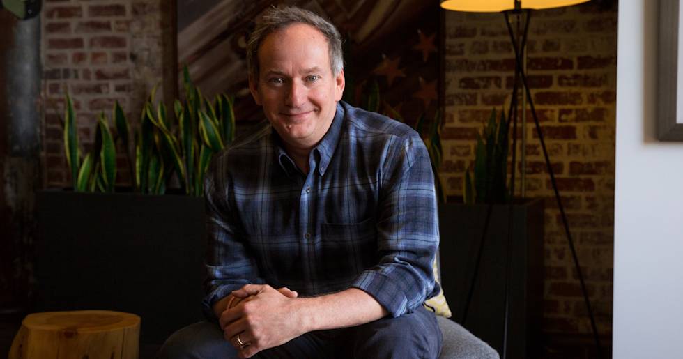 A man in a plaid button-down shirt sits casually on a chair, smiling warmly in a room with exposed brick walls and indoor plants