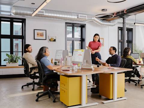 Office scene with four people collaborating at desks with computers, open-space design, and modern decor.