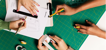 Hands working on architectural drawings and sketches on a green cutting mat surface with drafting tools and measuring equipment