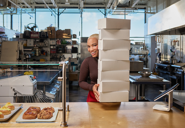 Restaurant worker in brown top and red apron smiling while carrying tall stack of white takeout boxes in commercial kitchen