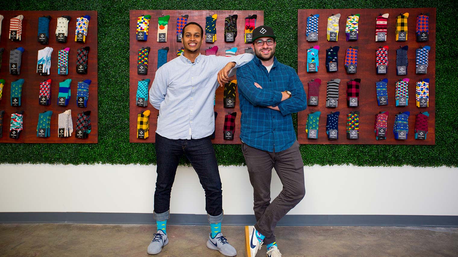 Stefan Lewinger and Futhum Tewolde of Sock Fancy smiling in front of a wall covered in socks. 