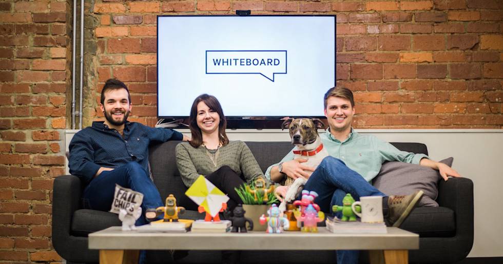 Three team members and a dog relaxing on a gray couch in an office with exposed brick walls and colorful toys on the coffee table
