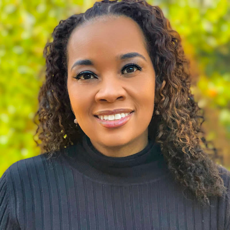 Professional headshot against blurred green foliage background, featuring warm smile and natural curly hair, wearing black sweater