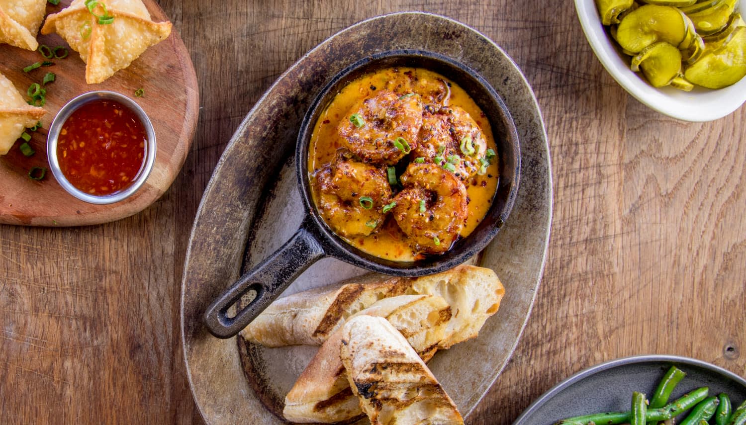 Table set with food, including shrimp and grits with bread, green beans, wontons with chili sauce, and pickles