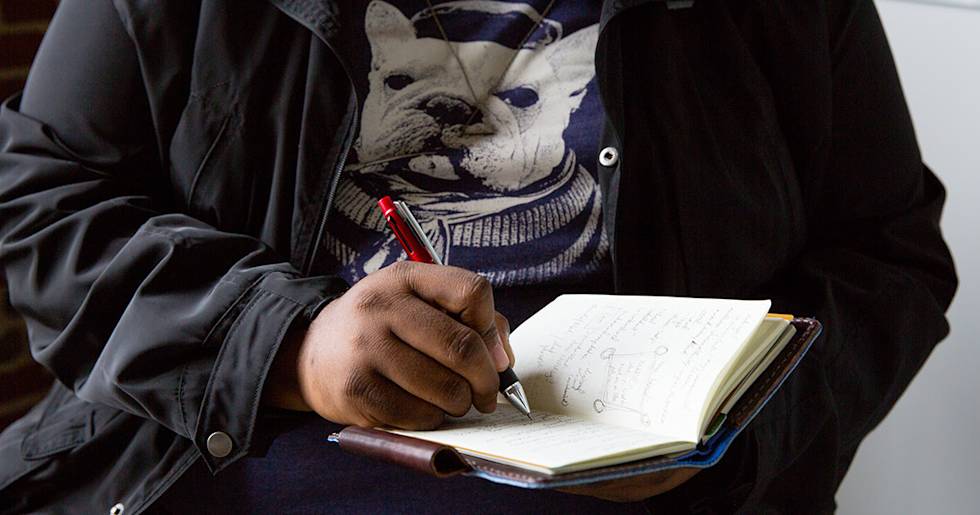 Person in black jacket with French bulldog graphic t-shirt writing in small leather notebook with red pen