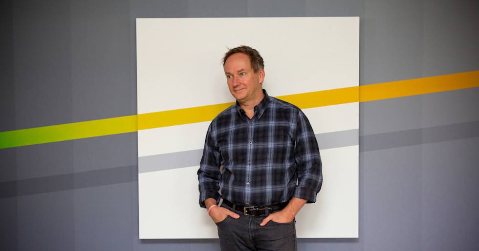 Professional in plaid button-down shirt and jeans stands against white backdrop with diagonal yellow and green stripes on gray wall