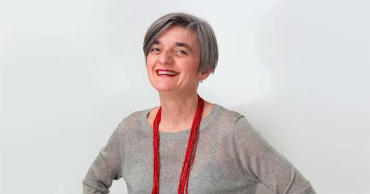 Professional headshot of Alessandra Farabegoli with short gray hair wearing a gray sweater and long red beaded necklace, smiling warmly against white background