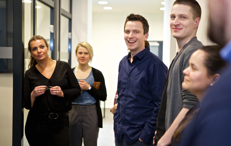 A group of young professionals standing in a bright office hallway, smiling and engaged in casual conversation
