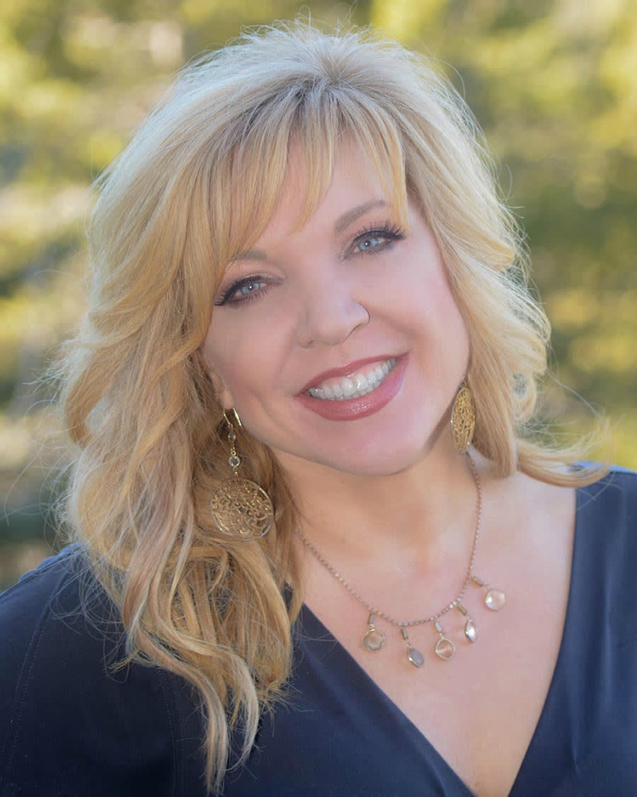 Professional headshot of Allison Crumpton with blonde hair wearing a navy top, gold earrings and delicate necklace, smiling warmly against blurred greenery