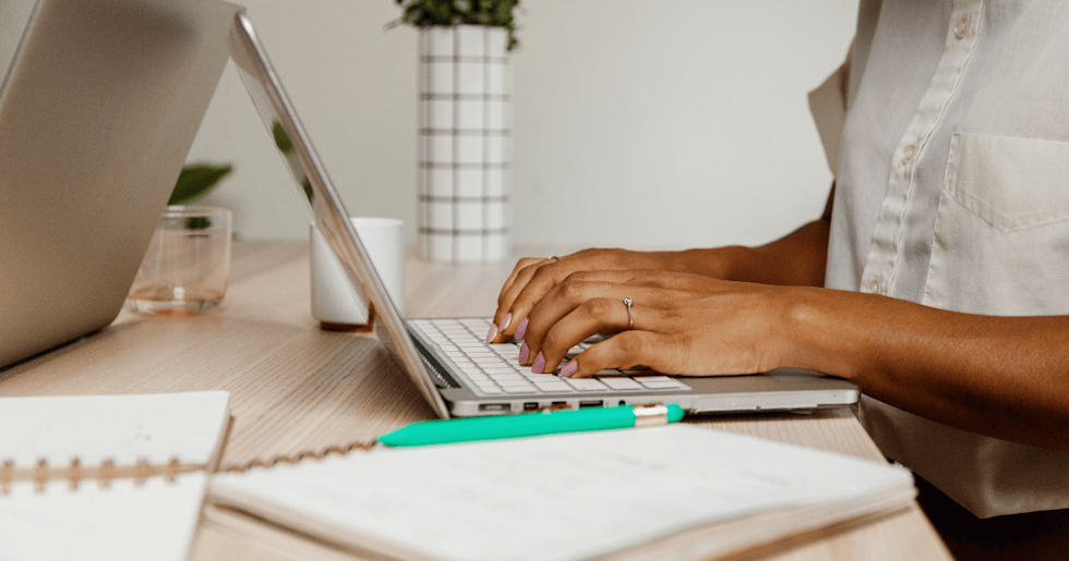 A person sits at a light-colored desk, typing on a laptop while taking notes in a nearby notebook with a pen.