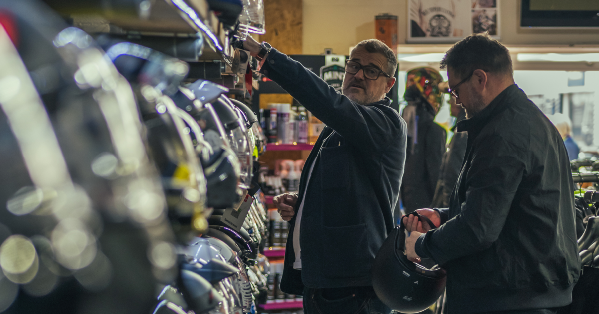 Two men are browsing helmets in a motorcycle shop, both wearing dark jackets and eyeglasses. One man is reaching toward a shelf with a variety of motorcycle helmets. The other man is holding a black helmet.