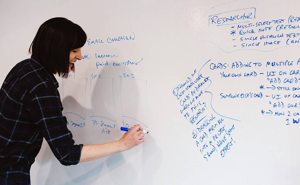 Person in black plaid shirt writing on whiteboard with blue marker, showing project notes and diagrams