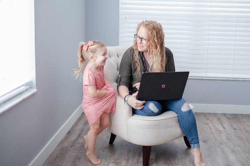 Rachel Parrish and a young child share a joyful moment while looking at laptop together on a beige armchair in bright room