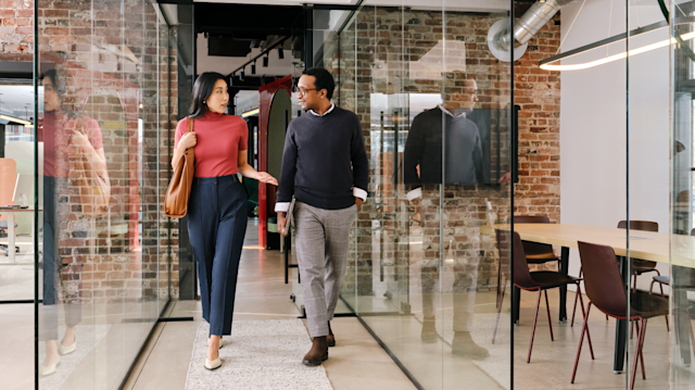 Two people walk together in a modern office hallway with glass walls and brick accents, engaging in a conversation.