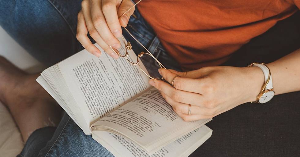 Hands holding reading glasses over open book pages, person wearing orange shirt, jeans, and white watch with gold details