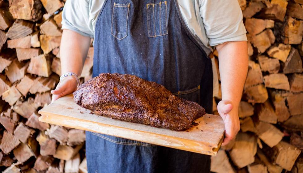 Man in a blue apron holding a large cut of barbecued meat while standing in front of a stack of firewood