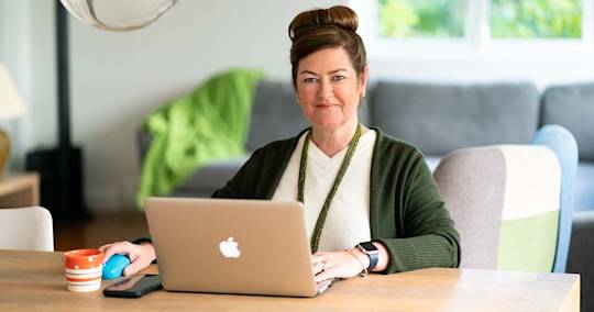 Professional woman in green cardigan working at wooden desk with MacBook laptop, coffee mug, and modern home office setting