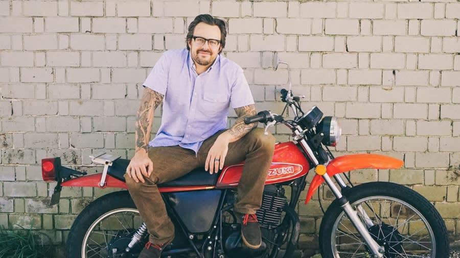A person in a light purple shirt and brown pants sits casually on a vintage red Suzuki motorcycle against a brick wall