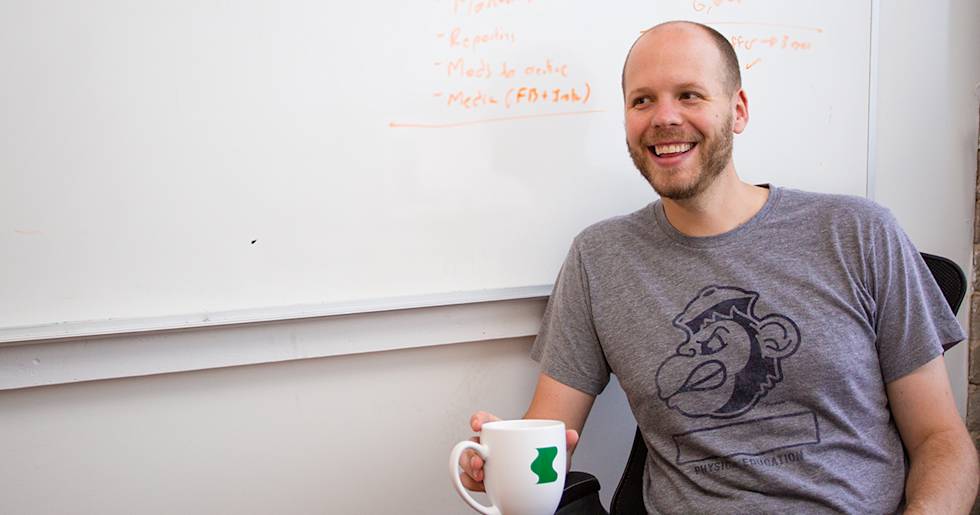 A smiling professional in a gray t-shirt with monkey graphic sits by whiteboard, holding white coffee mug with green logo