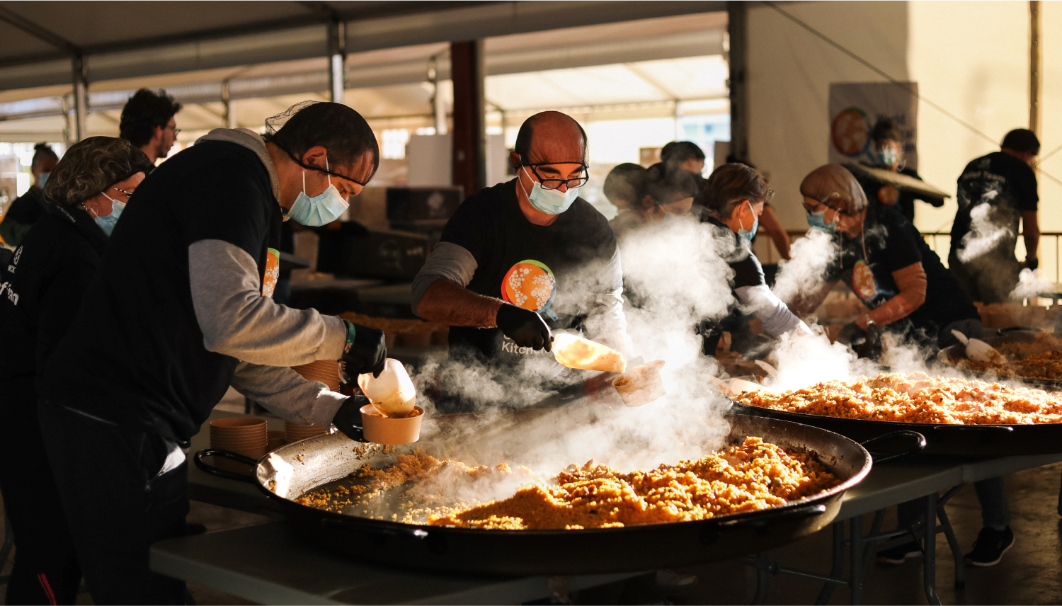 Chefs wearing World Central Kitchen shirts in a large tent scooping food out of large pans into single-serving-size bowls.
