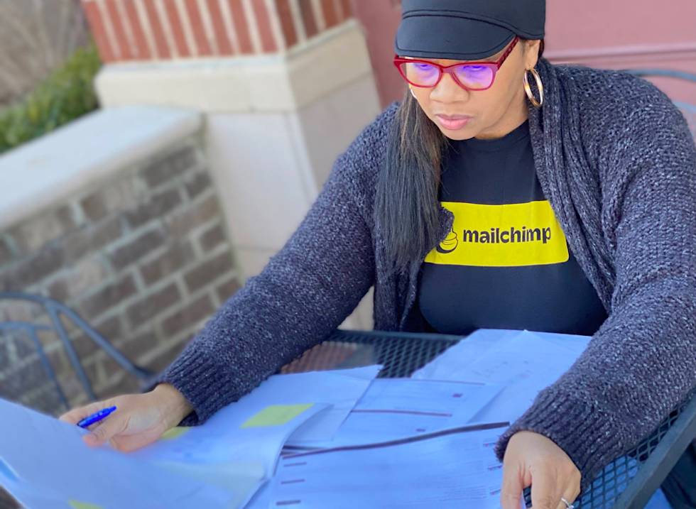 Person wearing Mailchimp t-shirt and red glasses reviews documents while seated outdoors at metal table, dressed in navy cap and gray cardigan