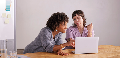 Two colleagues collaborating at a wooden desk, looking at a laptop screen while having a casual discussion in an office setting