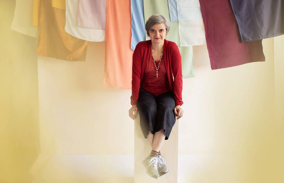 A photograph of a woman sitting on a stool in front of colorful linens.